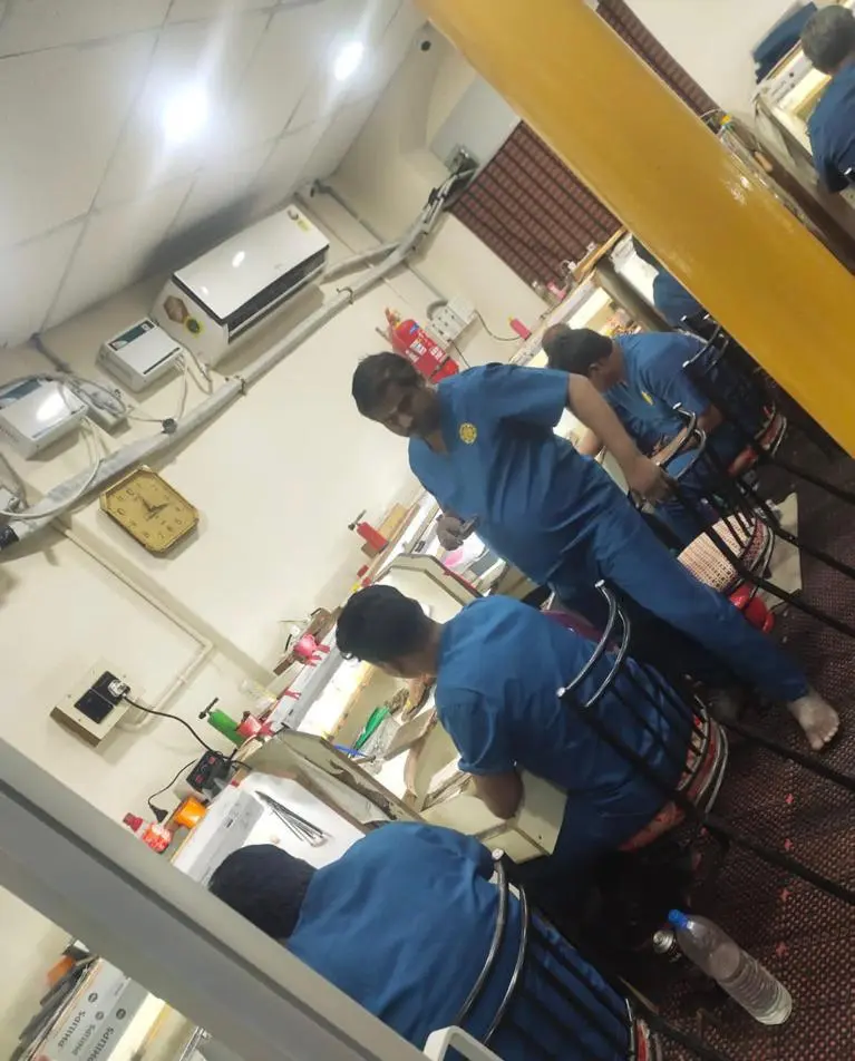 Group of workers in blue scrubs seated at a long workstation in a busy workshop, with tools and equipment nearby.