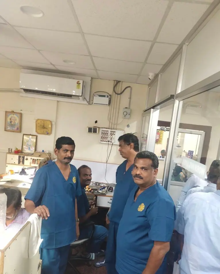 Four men in blue scrubs stand in a clinic room with beige walls and equipment, likely medical staff taking a break or chatting.