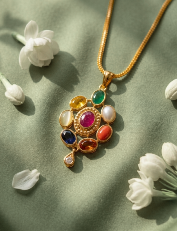 Gold pendant necklace featuring a cluster of multicolored gemstones on a gold chain, set on soft sage-green fabric with white flowers nearby.