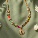 Gold necklace with flower-shaped diamond clusters and multicolored gems on a green fabric backdrop, pendant featuring a green teardrop emerald.