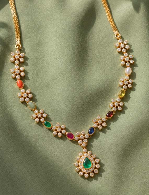 Gold necklace with flower-shaped diamond clusters and multicolored gems on a green fabric backdrop, pendant featuring a green teardrop emerald.