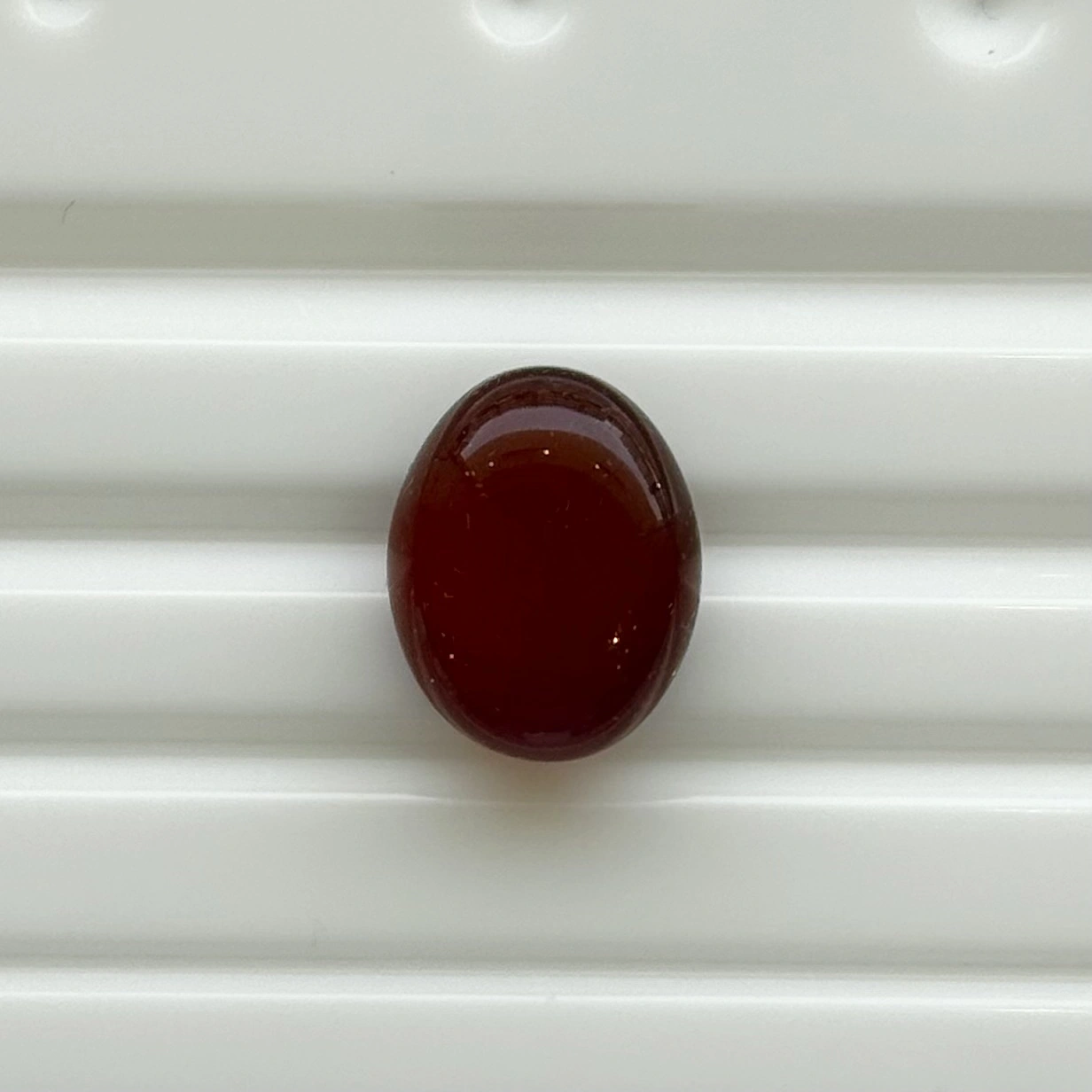 Aqeeq_192-39748 - N Gopaldaas Jewellers A dark red oval bead resting on a white ribbed surface.