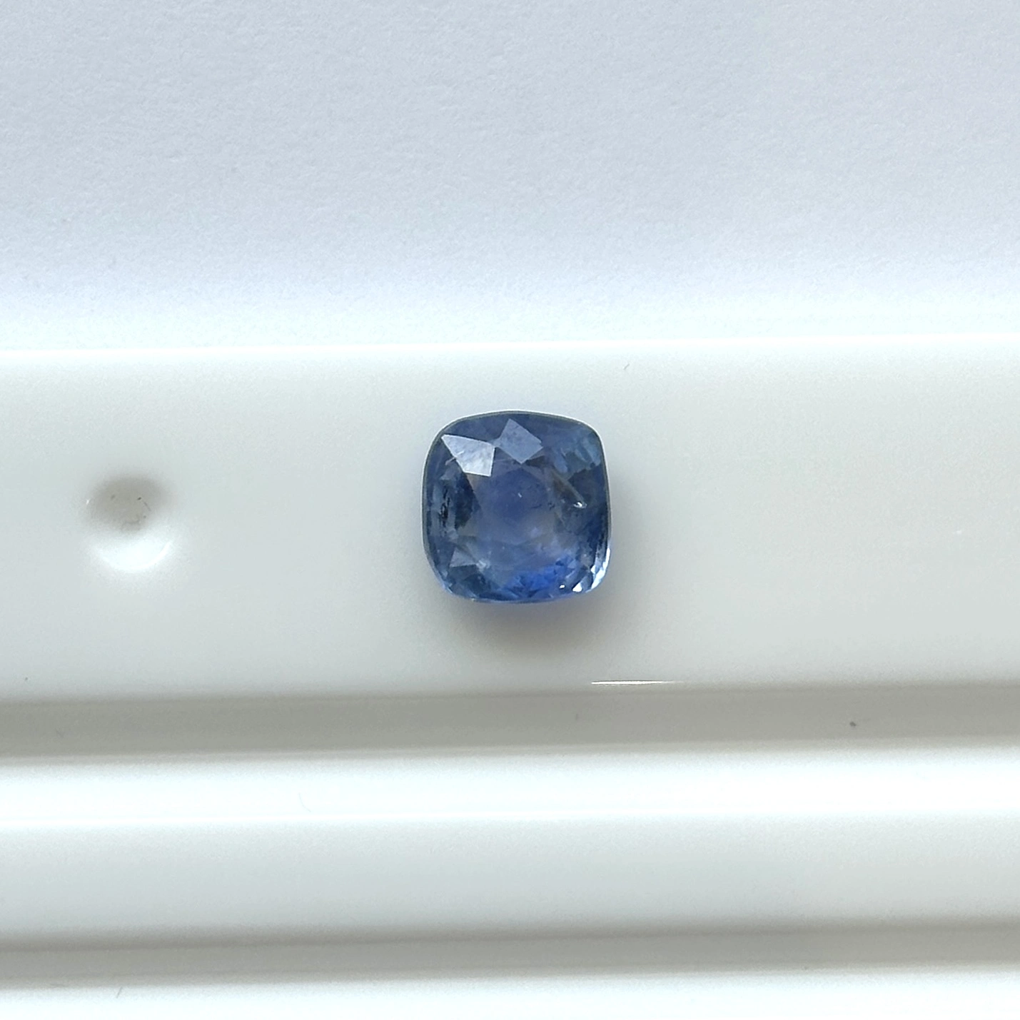 blue sapphire_190-34779 - N Gopaldaas Jewellers A cushion-cut blue gemstone placed on a white surface, showcasing its faceted facets and rich color.